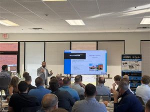 Conference room presentation on ONICON flow and energy measurement solutions. Presenter explains company expertise and technologies shown on a large screen, with ONICON branding visible on a banner