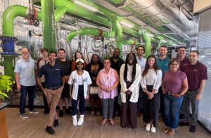 Group photo of new hires standing in front of an industrial-themed mural featuring pipes, ducts, and valves. Taken indoors in a modern office space during ONICON’s June 2025 new hire lunch event.